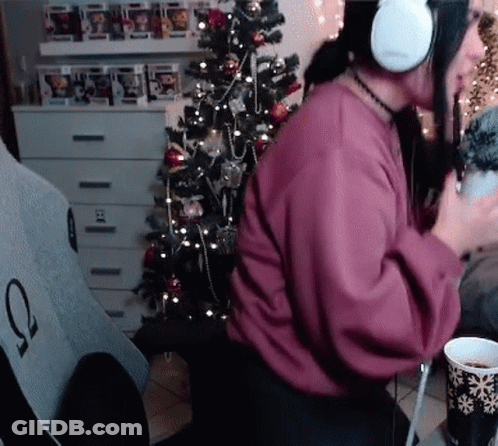 A Woman Wearing Headphones Is Sitting In Front Of A Christmas Tree Holding A Cup Of Coffee . GIF