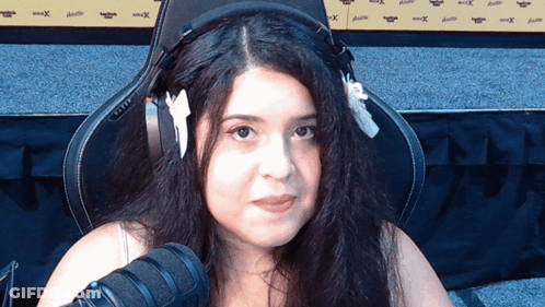 A Woman Wearing Headphones Sits In Front Of A Microphone And A Wall That Says Xtreme GIF