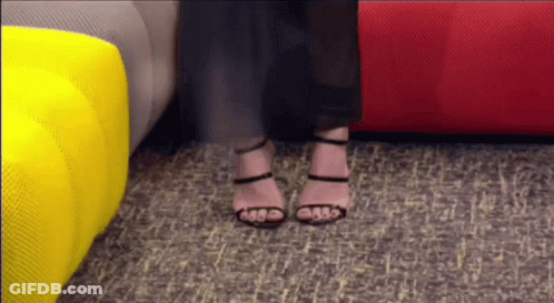 A Woman Wearing High Heels Is Standing In Front Of A Red Couch GIF