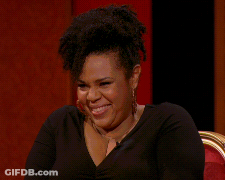 A Woman Wearing Hoop Earrings And A Black Shirt Is Laughing While Sitting In A Chair . GIF