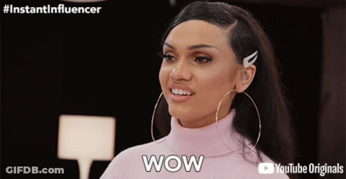 A Woman Wearing Hoop Earrings And A Pink Turtleneck Sweater Is Saying Wow GIF