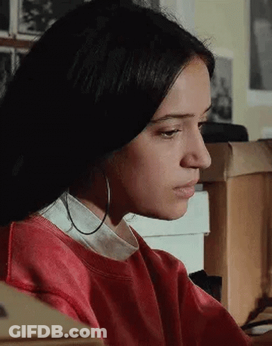A Woman Wearing Hoop Earrings And A Red Sweater Is Sitting At A Desk . GIF