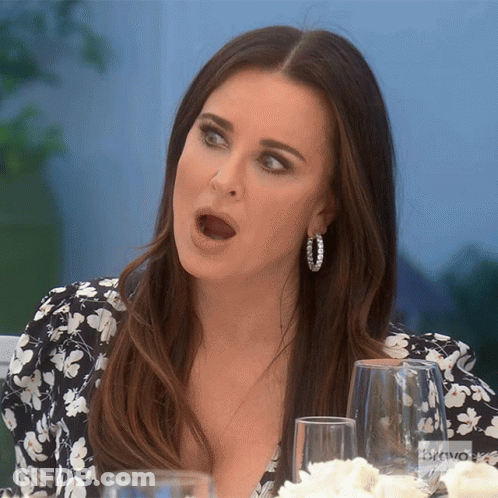 A Woman Wearing Hoop Earrings Is Sitting At A Table With A Bravo Logo In The Background GIF