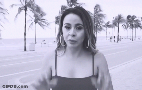 A Woman Wearing Hoop Earrings Stands In Front Of Palm Trees On The Beach GIF