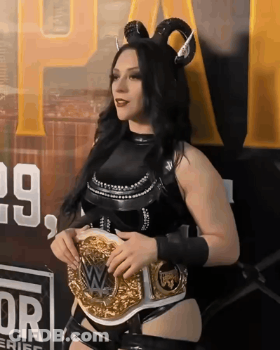 A Woman Wearing Horns And Holding A Wrestling Belt In Front Of A Sign That Says 29 GIF
