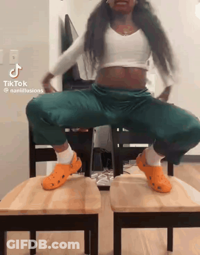 A Woman Wearing Orange Crocs Is Squatting On Two Wooden Chairs .. GIF
