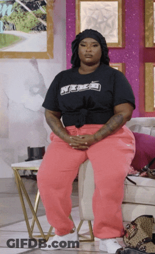A Woman Wearing Pink Sweatpants And A Black Shirt That Says ' A ' On It Sits In A Chair GIF