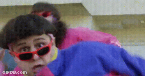 A Woman Wearing Red Sunglasses And A Pink Jacket Is Standing Next To Another Woman Wearing A Blue Jacket . GIF