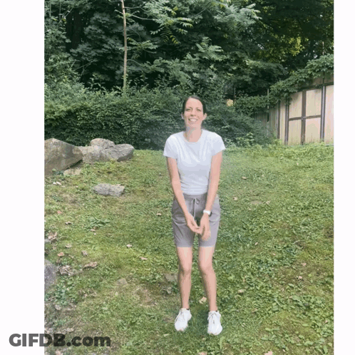 A Woman Wearing Shorts And A White Shirt Stands In A Grassy Field GIF