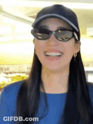 A Woman Wearing Sunglasses And A Baseball Cap Smiles For The Camera GIF