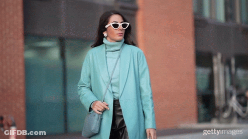 A Woman Wearing Sunglasses And A Blue Coat Is Walking Down The Street GIF