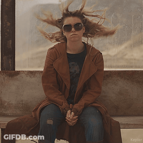 A Woman Wearing Sunglasses And A Brown Coat Sits On A Bench In Front Of A Window That Says Kepler On It GIF