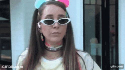 A Woman Wearing Sunglasses And A Colorful Headband Is Standing In Front Of A Building . GIF