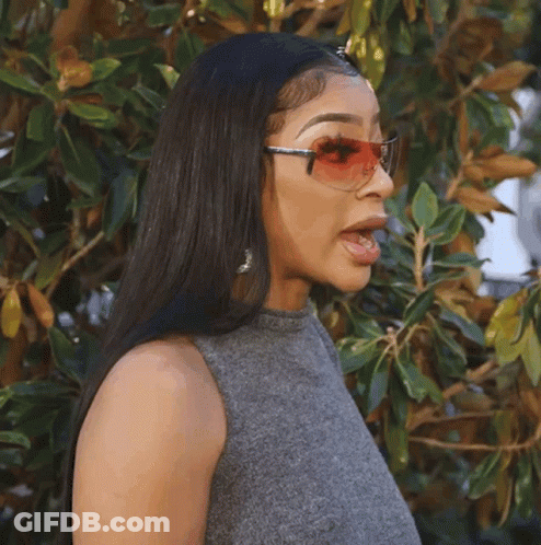 A Woman Wearing Sunglasses And A Gray Top Stands In Front Of A Tree GIF