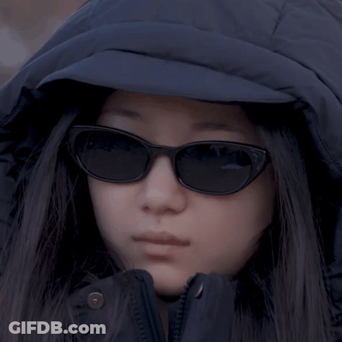 A Woman Wearing Sunglasses And A Hooded Jacket Looks At The Camera GIF