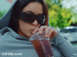 A Woman Wearing Sunglasses And A Hoodie Drinks From A Plastic Cup GIF