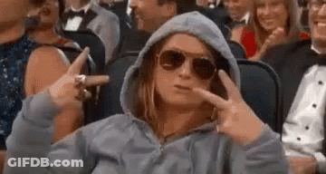 A Woman Wearing Sunglasses And A Hoodie Is Sitting In A Crowd Of People And Making A Peace Sign . GIF