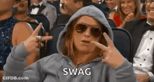 A Woman Wearing Sunglasses And A Hoodie Is Sitting In A Crowd Of People And Making A Swag Sign . GIF