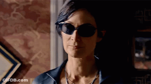 A Woman Wearing Sunglasses And A Necklace Is Standing In A Room . GIF