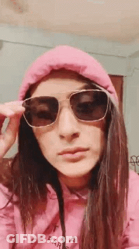 A Woman Wearing Sunglasses And A Pink Hoodie Is Taking Off Her Sunglasses . GIF