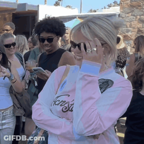 A Woman Wearing Sunglasses And A Pink Shirt That Says Fang Sits In A Crowd GIF