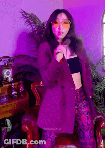 A Woman Wearing Sunglasses And A Purple Jacket Stands In Front Of A Purple Wall GIF