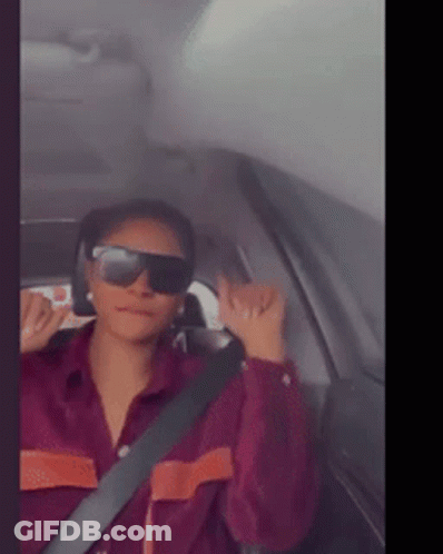 A Woman Wearing Sunglasses And A Purple Shirt Is Sitting In A Car GIF