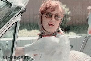 A Woman Wearing Sunglasses And A Scarf Is Driving A White Car . GIF