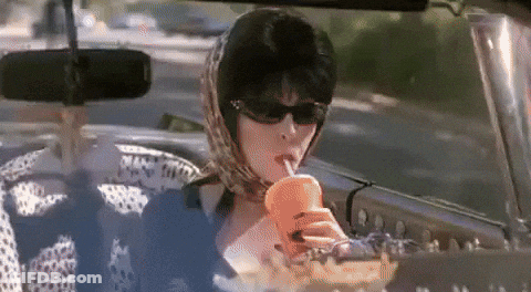 A Woman Wearing Sunglasses And A Scarf Is Sitting In A Car Drinking From A Cup Through A Straw . GIF