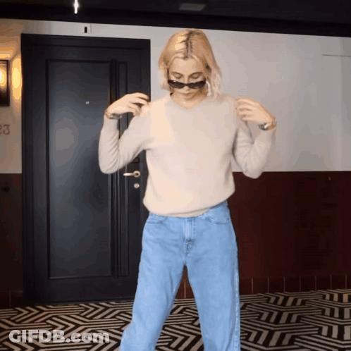 A Woman Wearing Sunglasses And A Sweater Is Standing In Front Of A Door That Has The Number 23 On It GIF
