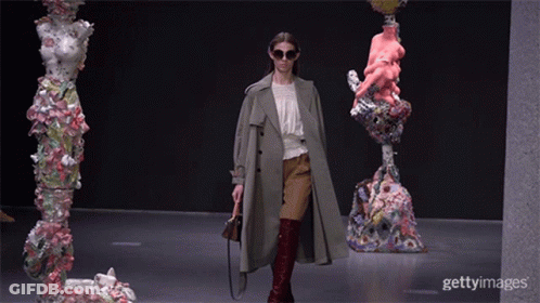 A Woman Wearing Sunglasses And A Trench Coat Walks Down A Runway In Front Of Sculptures GIF