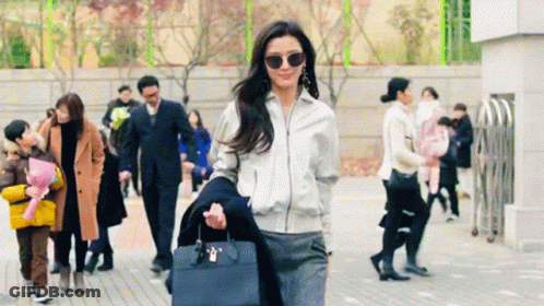 A Woman Wearing Sunglasses And A White Jacket Is Walking Down A Sidewalk GIF