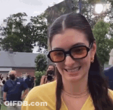 A Woman Wearing Sunglasses And A Yellow Shirt Is Smiling And Looking At The Camera . GIF