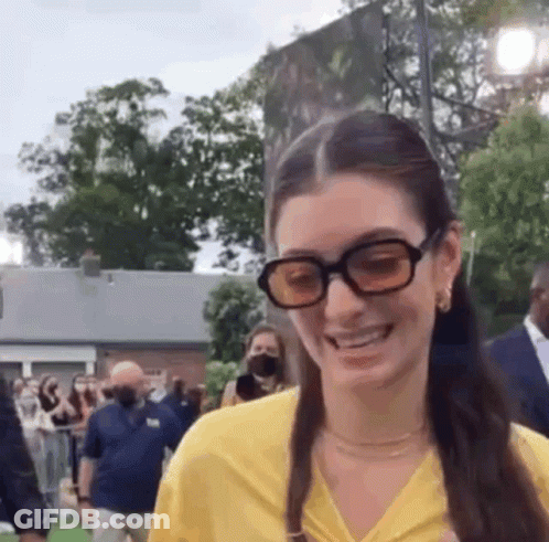 A Woman Wearing Sunglasses And A Yellow Shirt Is Smiling In Front Of A Crowd Of People . GIF