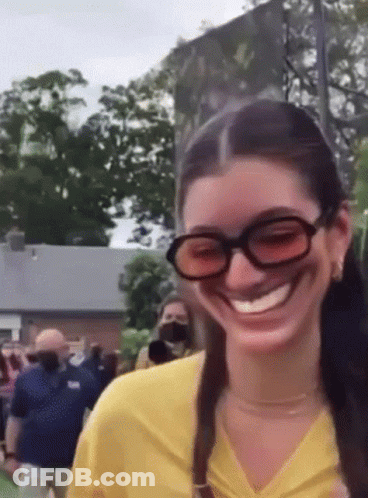 A Woman Wearing Sunglasses And A Yellow Shirt Is Smiling While Standing In Front Of A Crowd . GIF