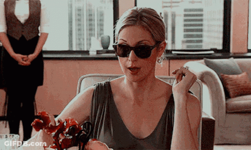 A Woman Wearing Sunglasses And Earrings Sits At A Table GIF