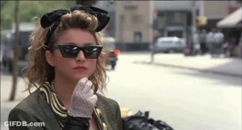 A Woman Wearing Sunglasses And Gloves Is Standing On A City Street GIF