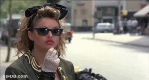 A Woman Wearing Sunglasses And Gloves Is Standing On A Street . GIF