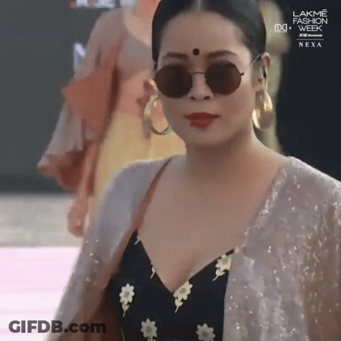 A Woman Wearing Sunglasses And Hoop Earrings Is Walking Down A Runway At Lakme Fashion Week GIF