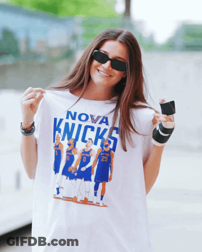 A Woman Wearing Sunglasses Holds Up A Nova Knicks Shirt GIF