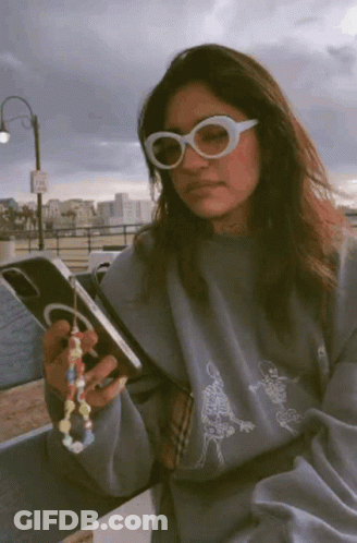 A Woman Wearing Sunglasses Is Holding A Cell Phone In Her Hand GIF