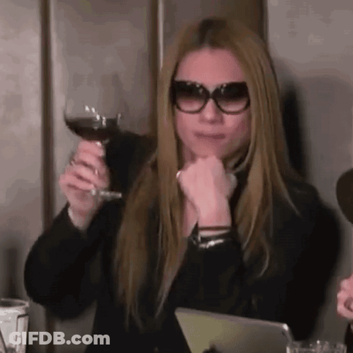 A Woman Wearing Sunglasses Is Sitting At A Table Holding A Glass Of Red Wine . GIF