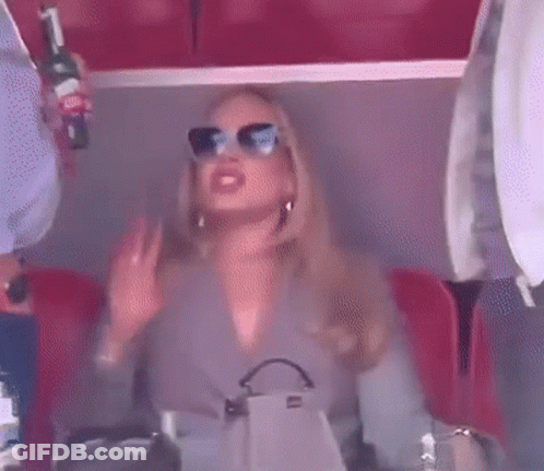 A Woman Wearing Sunglasses Is Sitting In A Stadium Holding A Bottle Of Wine And A Purse . GIF