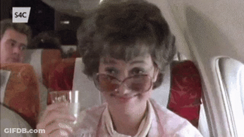 A Woman Wearing Sunglasses Is Sitting On An Airplane Holding A Glass GIF