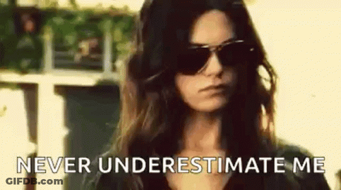 A Woman Wearing Sunglasses Is Standing In Front Of A Building And Saying `` Never Underestimate Me '' . GIF