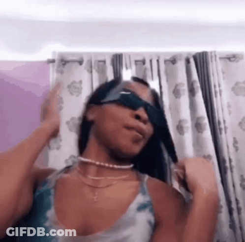 A Woman Wearing Sunglasses Is Standing In Front Of A Curtain . GIF