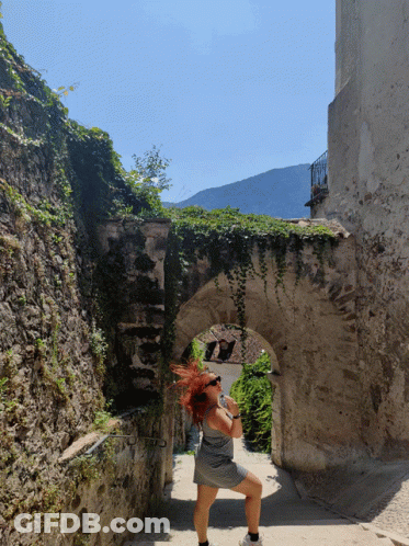A Woman Wearing Sunglasses Stands In A Stone Archway GIF