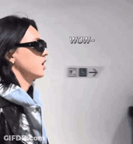 A Woman Wearing Sunglasses Stands In Front Of A Sign That Says Wow On It GIF