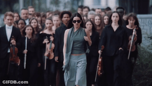 A Woman Wearing Sunglasses Walks In Front Of A Large Group Of Musicians GIF