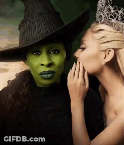 A Woman Whispering Into Another Woman 'S Ear While Wearing A Witch Costume GIF